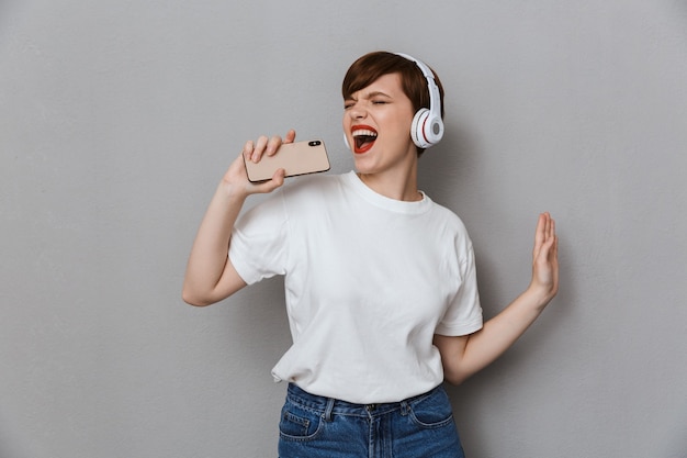 Retrato de uma jovem feliz cantando enquanto ouve música com fones de ouvido e smartphone isolados sobre uma parede cinza