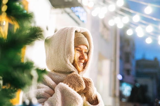 Retrato de uma jovem feliz com cabelo encaracolado em casaco de pele se divertindo na rua de inverno decorada com luzes