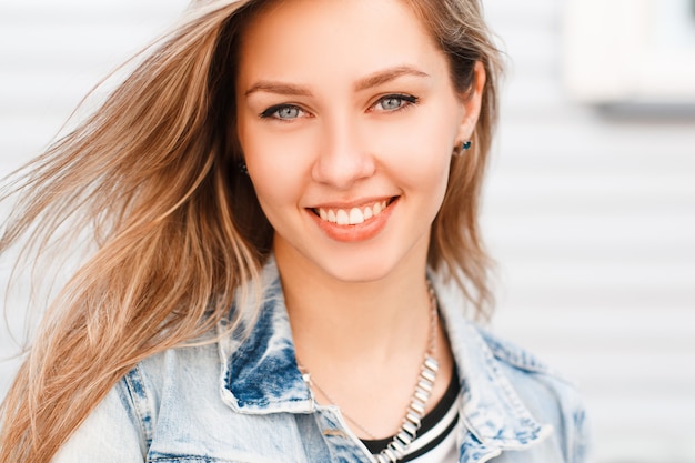 Retrato de uma jovem feliz, com incríveis olhos azuis com um sorriso maravilhoso com maquiagem natural em uma jaqueta jeans da moda em uma camiseta em uma parede branca em um dia ensolarado de verão. garota alegre.