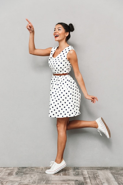 Retrato de uma jovem feliz com vestido de verão isolado, posando em pé, apontando