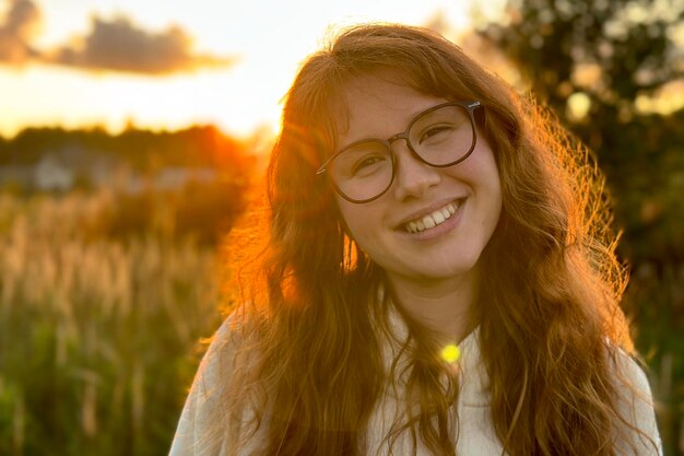 Retrato de uma jovem feliz de óculos ao pôr do sol no fundo natural, sorrindo e olhando para a câmera