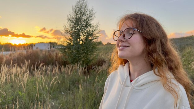 Retrato de uma jovem feliz de óculos ao pôr do sol no fundo natural, sorrindo e olhando para a câmera