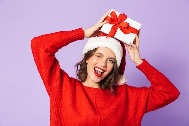 Retrato de uma jovem feliz e animada com chapéu de natal, isolado na parede roxa, segurando uma caixa de presente surpresa.