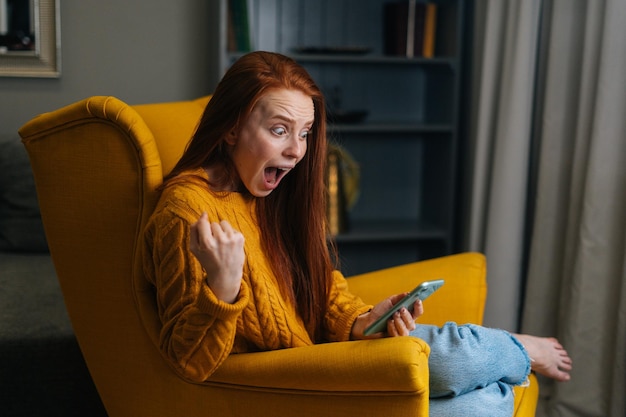 Retrato de uma jovem feliz e excitada olhando a tela do celular sentada em uma poltrona amarela rindo com boas notícias sentindo uma grande surpresa positiva