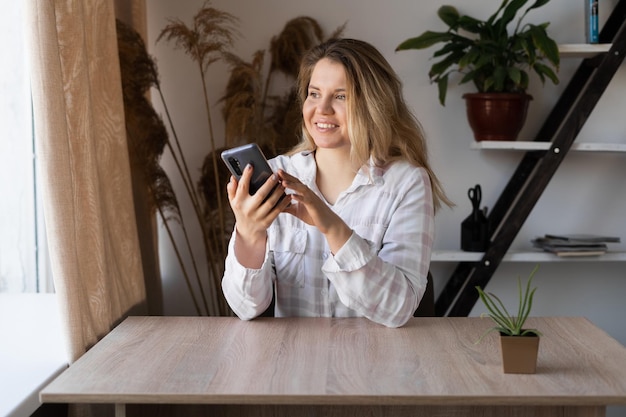 Retrato de uma jovem feliz e positiva que lê um SMS em um smartphone e se senta sorrindo pela janela Garota no escritório com uma prateleira com flores e uma mesa