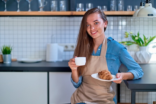 Retrato de uma jovem feliz e sorridente padeiro segurando um croissant de xícara e prato branco na cozinha do loft