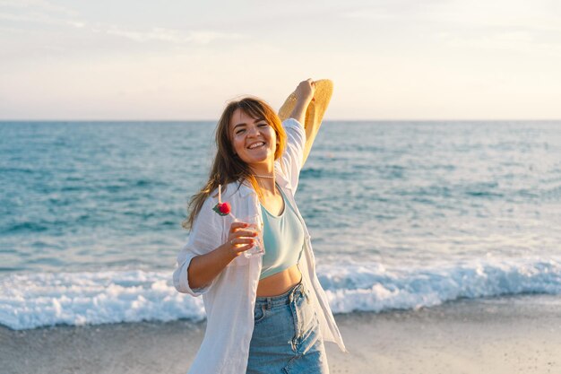 Retrato de uma jovem feliz em um chapéu de palha com um coquetel na mão em um fundo de mar