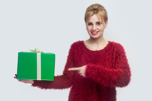 Retrato de uma jovem feliz na blusa vermelha em pé e segurando a caixa de presente verde e apontando o dedo, olhando para a câmera. Interior, isolado, tiro de estúdio, espaço de cópia, fundo cinza