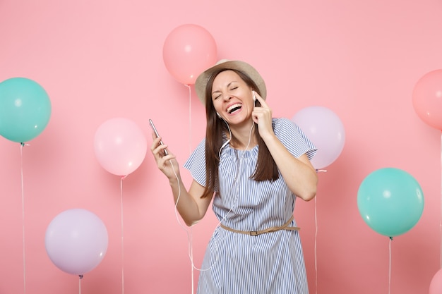 Retrato de uma jovem feliz rindo com chapéu de palha de verão e vestido azul com telefone celular e fones de ouvido, ouvindo música no fundo rosa pastel com balões de ar coloridos. Festa de aniversário.