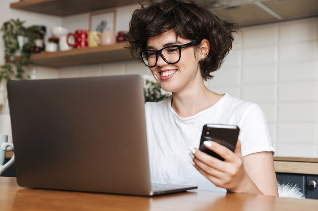 Retrato de uma jovem feliz usando óculos e trabalhando no laptop em casa pela manhã, segurando o telefone celular