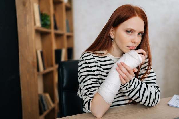 Retrato de uma jovem ferida pela dor com a mão direita quebrada envolta em bandagem de gesso branco sentada à mesa olhando para a câmera