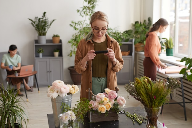 Retrato de uma jovem florista tirando fotos de composições florais no smartphone enquanto trabalha em uma floricultura, copie o espaço