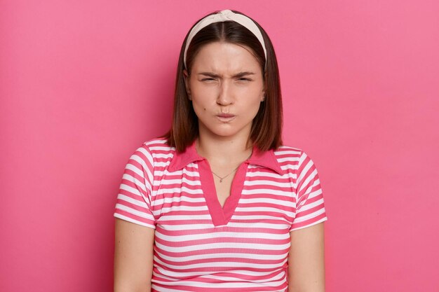 Retrato de uma jovem frustrada e zangada com cabelos castanhos, vestindo camiseta casual listrada em pé, fundo rosa isolado, dentes cerrados e olhos fechados, discutindo expressando emoções negativas e ódio