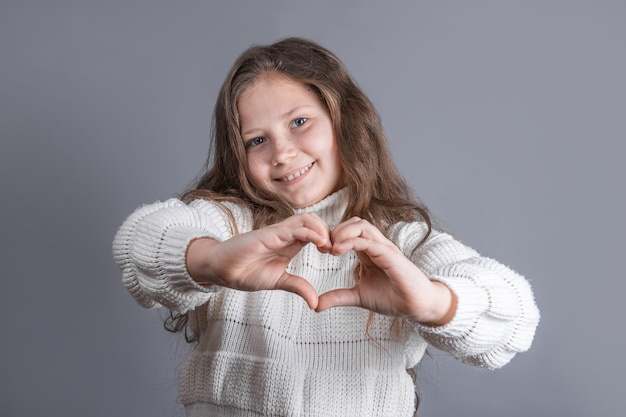 Retrato de uma jovem garota atraente com cabelo longo esvoaçante loiro em uma camisola branca sorrindo mostra as mãos do coração em um fundo cinza do estúdio. Lugar para texto. Copie o espaço.