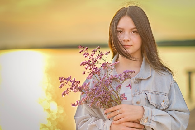 Retrato de uma jovem garota com um buquê de flores nas mãos contra o pôr do sol.