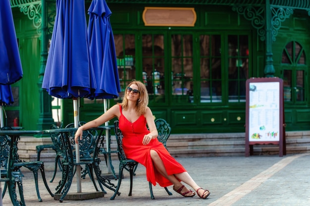 Retrato de uma jovem garota com um vestido vermelho brilhante, sentado à mesa em um café vazio de verão. Uma jovem em um restaurante. Para sentar sozinho. Mulher bonita em óculos de sol. Uma loira com um vestido vermelho.