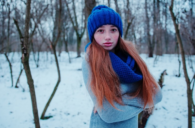 Retrato de uma jovem garota de cabelo vermelho com sardas vestindo no chapéu de malha de lã azul e lenço em dia de inverno.