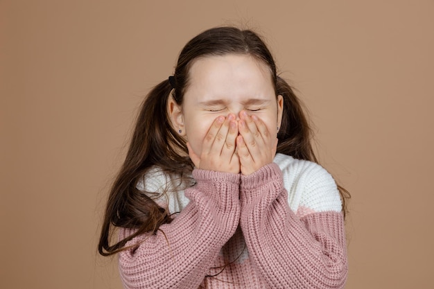 Retrato de uma jovem garota incrível com longos cabelos escuros no suéter rosa branco em pé com os olhos fechados estragando os olhos cobrindo a boca com as mãos no fundo marrom