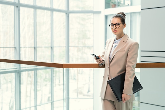 Retrato de uma jovem gerente séria em traje bege em pé com a pasta e procurando informações no smartphone