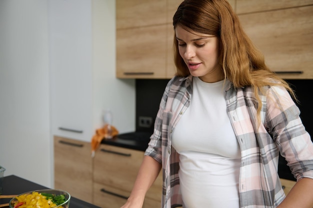 Retrato de uma jovem grávida cozinhando em casa preparando uma refeição vegetal saudável em pé na ilha da cozinha