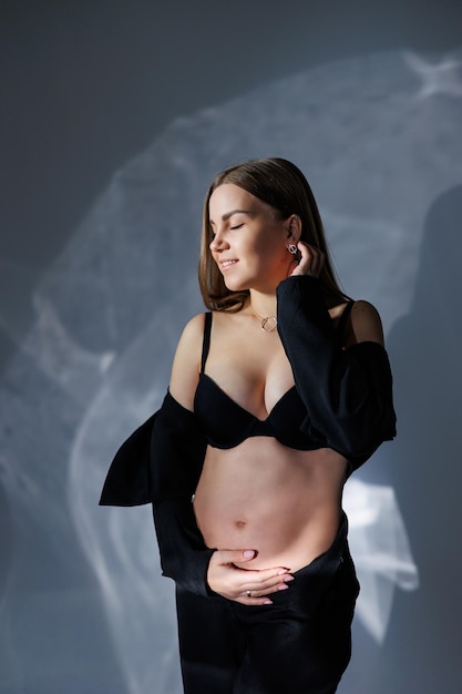 Retrato de uma jovem grávida em um terno clássico preto Uma mulher grávida em um fundo cinza abraça sua barriga