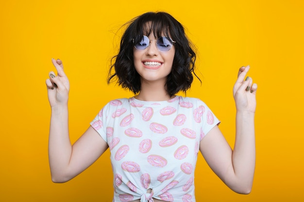 Retrato de uma jovem incrível com óculos em uma camisa com donuts apontando para cima com os olhos fechados, contra um fundo amarelo.