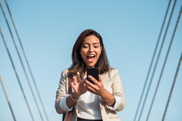 Retrato de uma jovem latina olhando animadamente para seu telefone celular inteligente na rua