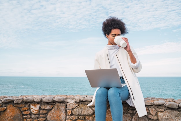 Retrato de uma jovem latina usando seu laptop e bebendo uma xícara de café enquanto está sentado contra o mar