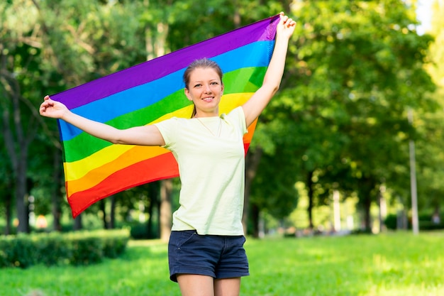 Retrato de uma jovem lésbica feliz orgulhosa, linda garota está acenando a bandeira gay de cor LGBT de arco-íris em um dia ensolarado de verão em fundo natural. Comunidade LGBT, conceito de igualdade de relações homossexuais