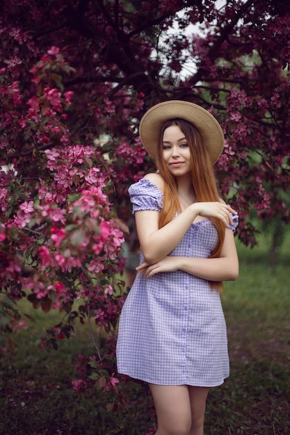 Retrato de uma jovem linda adolescente em um vestido roxo e em um chapéu fica ao lado de uma macieira rosa florescente