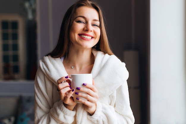 Retrato de uma jovem linda em um manto branco, desfrutando de um chá quente. Mulher segura uma xícara de chá ou café. Garota feliz sonhando.