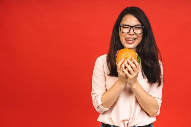 Retrato de uma jovem linda mulher faminta comendo hambúrguer retrato isolado de estudante com fast food sobre fundo vermelho conceito de dieta