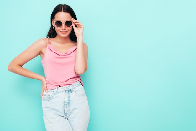 Retrato de uma jovem linda mulher sorridente em roupas da moda de verão Mulher despreocupada sexy posando perto da parede azul no estúdio Modelo positivo se divertindo dentro de casa Alegre e feliz em óculos de sol