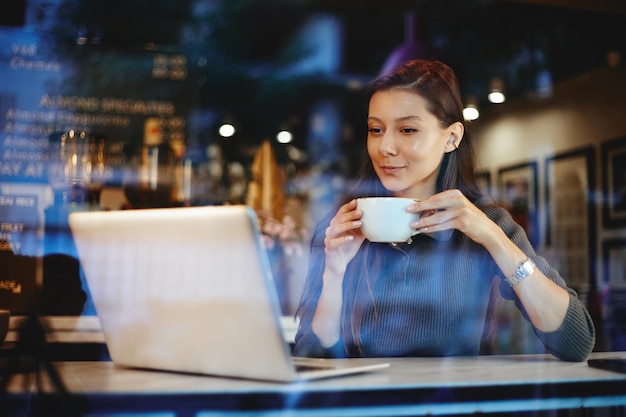 Retrato de uma jovem linda tomando café e usando o laptop em um café