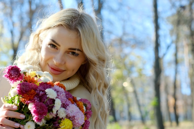 Retrato de uma jovem loira bonita com cabelos longos em um suéter branco e jaqueta em outono ensolarado em um parque da cidade com um buquê de flores