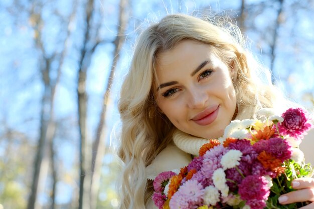 Retrato de uma jovem loira bonita com cabelos longos em um suéter branco e jaqueta em outono ensolarado em um parque da cidade com um buquê de flores