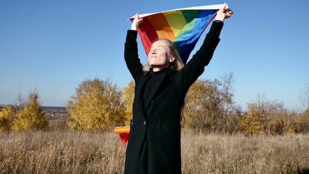 Retrato de uma jovem loira caucasiana com acne sem maquiagem segurando uma bandeira lgbt arco-íris no outono mulher de casaco preto em um dia ensolarado