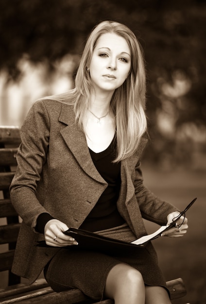 Retrato de uma jovem loira com roupa de negócios oficial, sentado no banco do parque com a leitura de documentos em um dia claro de verão. Conceito de bela empresária moderna