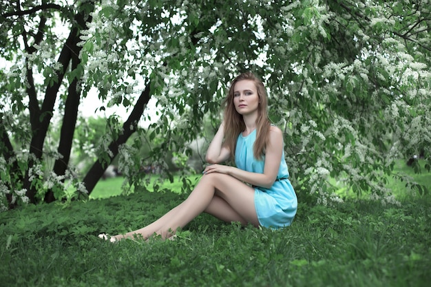 Retrato de uma jovem loira com um vestido azul. ela está sentada na grama. árvores em flores.