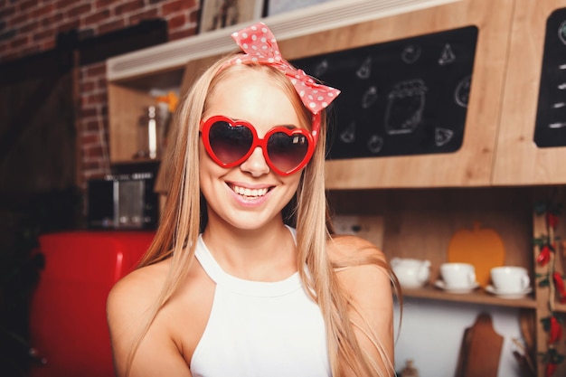 Retrato de uma jovem loira de cabelos longos usando óculos de sol olhando para a câmera e sorrindo na cozinha ensolarada confortável do loft vermelho