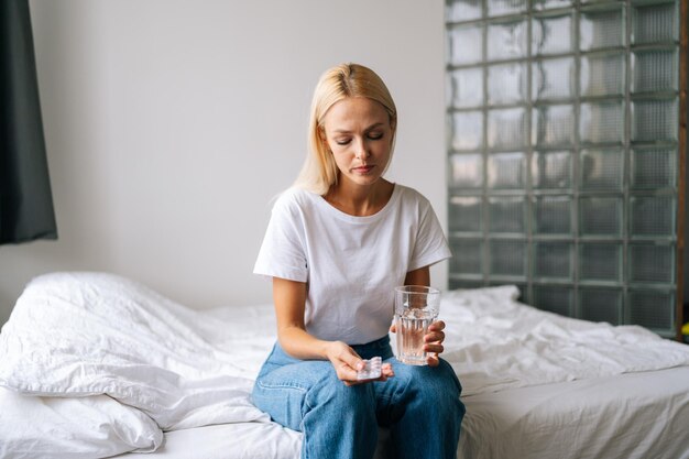 Retrato de uma jovem loira deprimida segurando comprimidos e copo de água nas mãos pensando em tomar drogas sentada na cama na sala de luz em casa