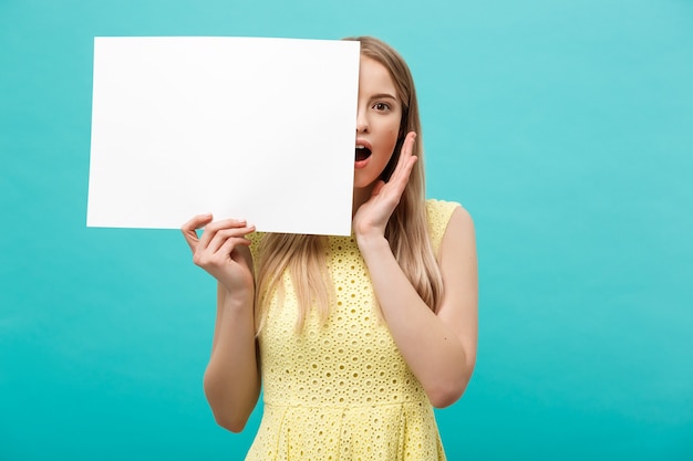 Retrato de uma jovem loira espantada, segurando um cartaz em branco com espaço de cópia no fundo azul do estúdio. mostrando rosto de surpresa chocado.