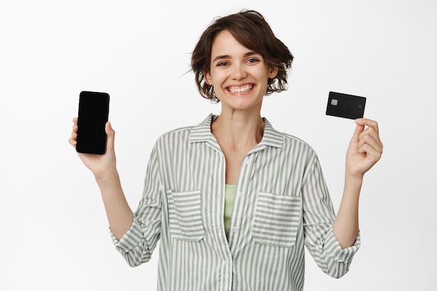 Foto retrato de uma jovem loira feliz mostrando o cartão de crédito plástico enquanto segura o celular isolado sobre fundo branco.