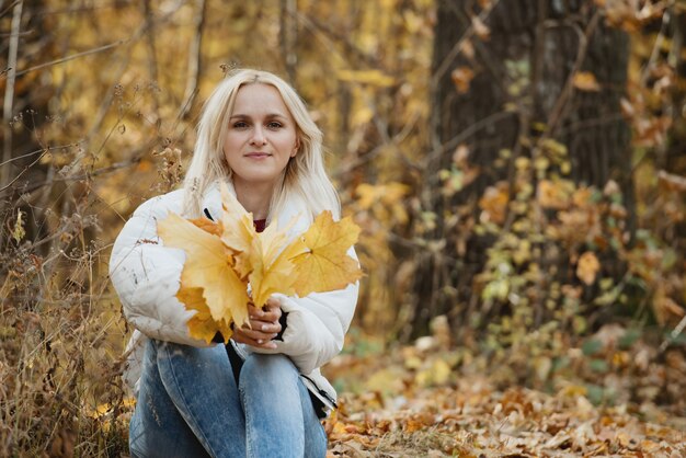 Retrato de uma jovem loira na floresta de outono, com um buquê de folhas amarelas nas mãos.