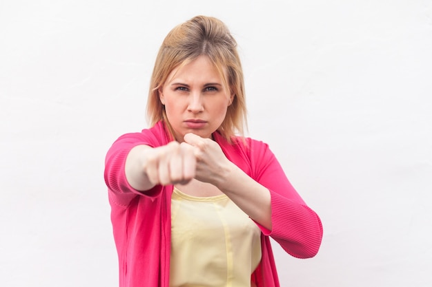 Retrato de uma jovem loira séria com raiva na camisa amarela e blusa vermelha em pé com os punhos de boxe, olhando e socando a câmera. tiro do estúdio interno, isolado no fundo da parede branca.