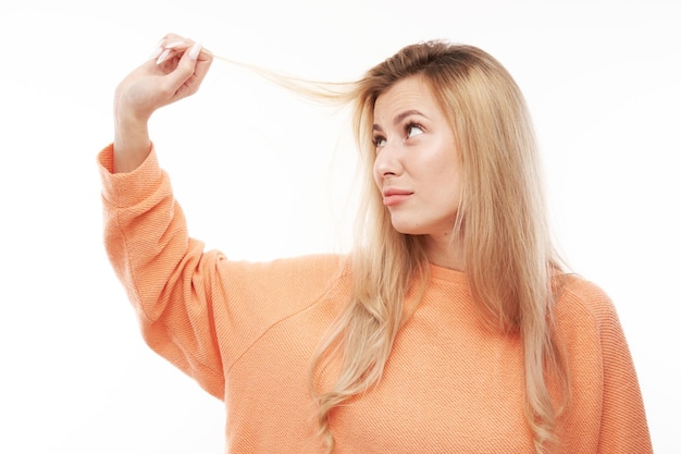 Foto retrato de uma jovem loira tocando o cabelo com rosto descontente isolado no fundo branco do estúdio conceito de cabelo danificado