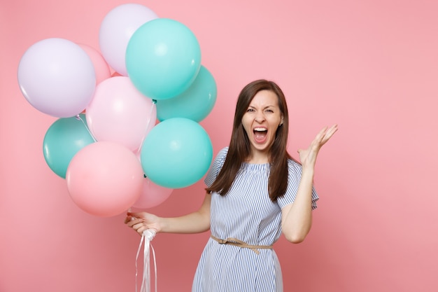 Retrato de uma jovem louca feliz num vestido azul gritando segurando balões de ar coloridos, espalhando as mãos isoladas no fundo rosa brilhante. Festa de aniversário, conceito de emoções sinceras de pessoas.