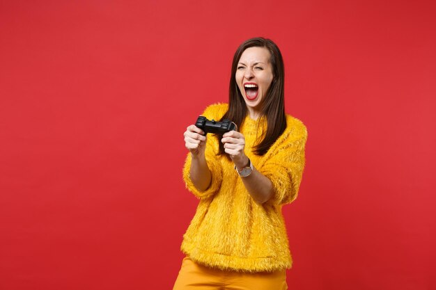 Retrato de uma jovem louca na camisola de pele amarela gritando, jogando videogame com joystick isolado no fundo da parede vermelha brilhante. emoções sinceras de pessoas, conceito de estilo de vida. simule o espaço da cópia.