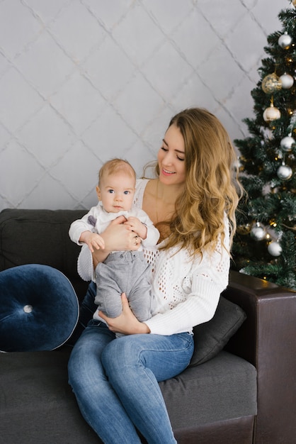 Retrato de uma jovem mãe em um suéter branco e seu jovem filho. eles estão sentados no sofá perto da árvore de natal