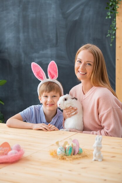 Retrato de uma jovem mãe feliz segurando um coelho fofo e sentado à mesa com o filho na orelha do coelho.